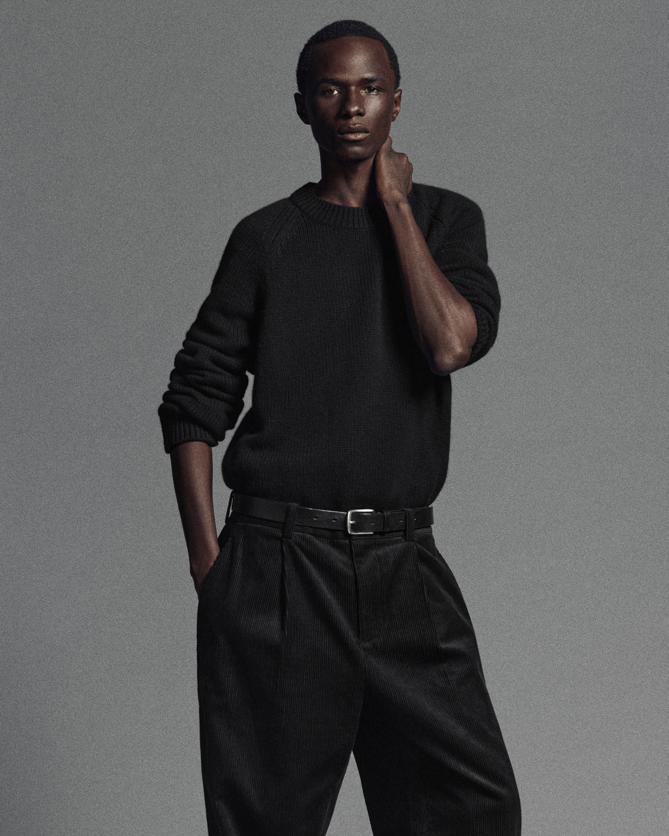 A person wearing a dark sweater and pleated pants stands with one hand in the pocket and the other touching their neck against a gray background.