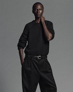Placeholder for: A person wearing a dark sweater and pleated pants stands with one hand in the pocket and the other touching their neck against a gray background.