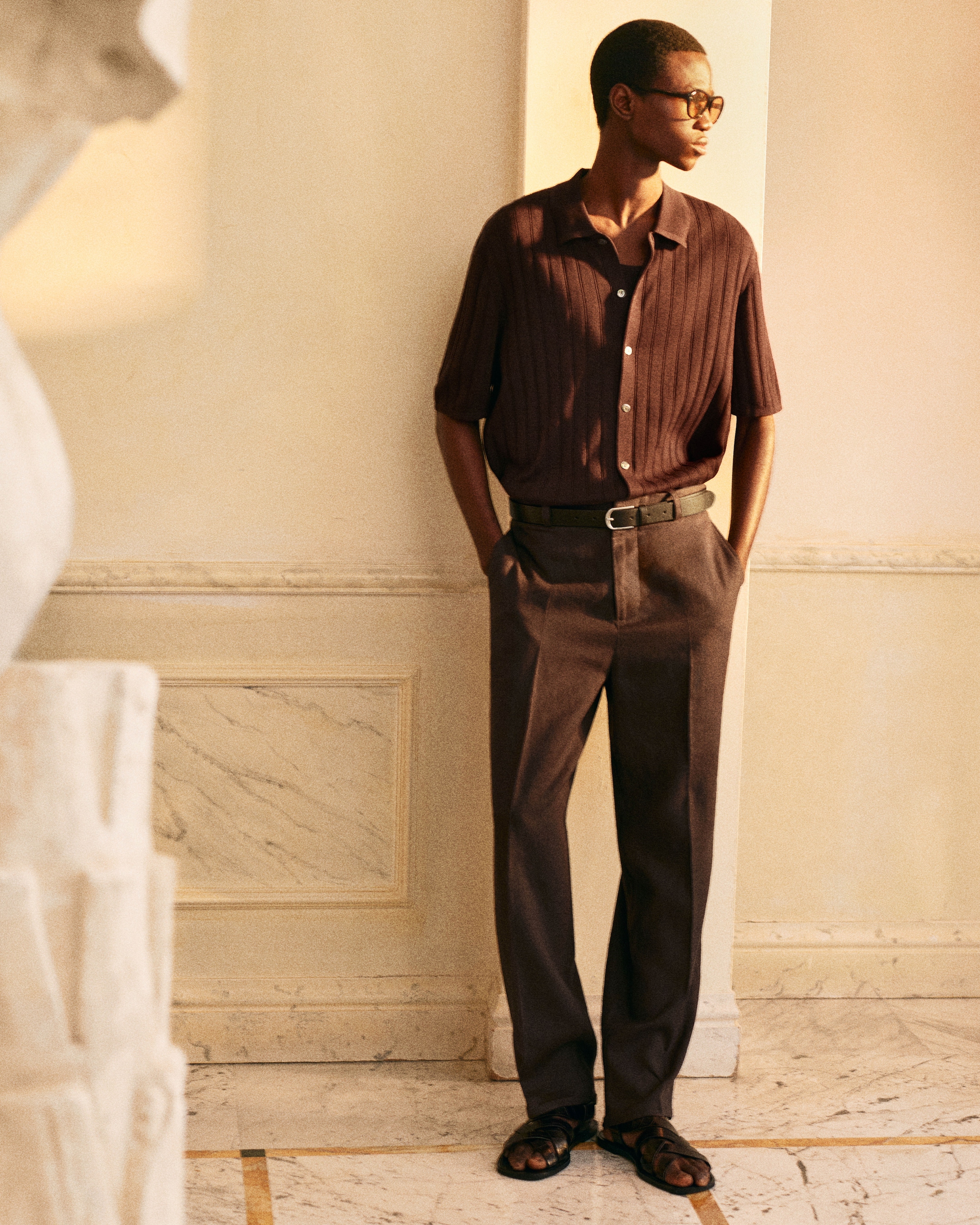 Man in a brown button-up shirt, trousers, and sandals, standing in a sunlit room with marble flooring, gazing to the side.