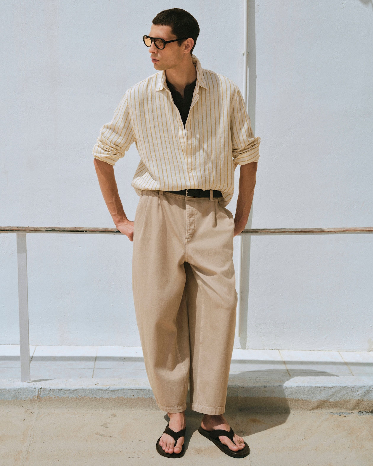 Man wearing sunglasses, a striped shirt, beige pants, and flip-flops stands casually against a white wall, hands in pockets.