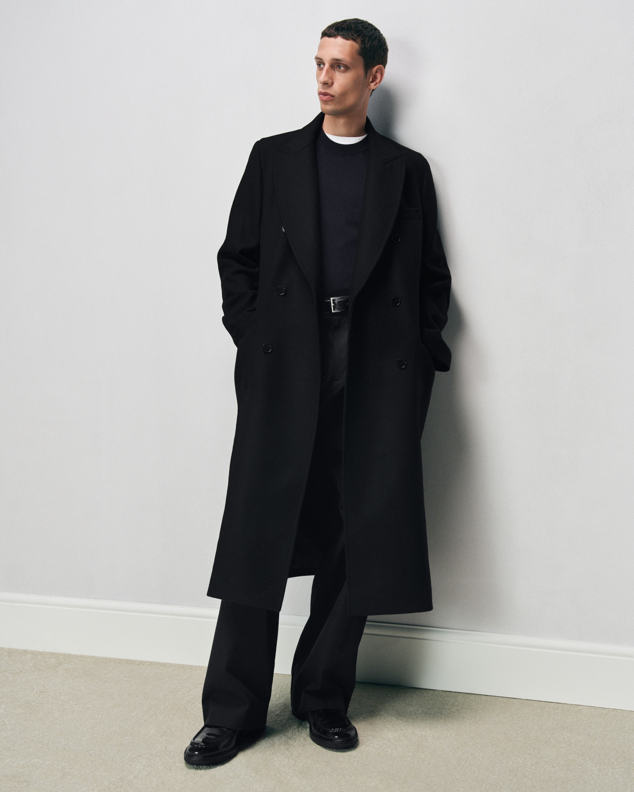 Person in a long black coat and black pants, standing against a plain white wall, hands in pockets, looking to the side.