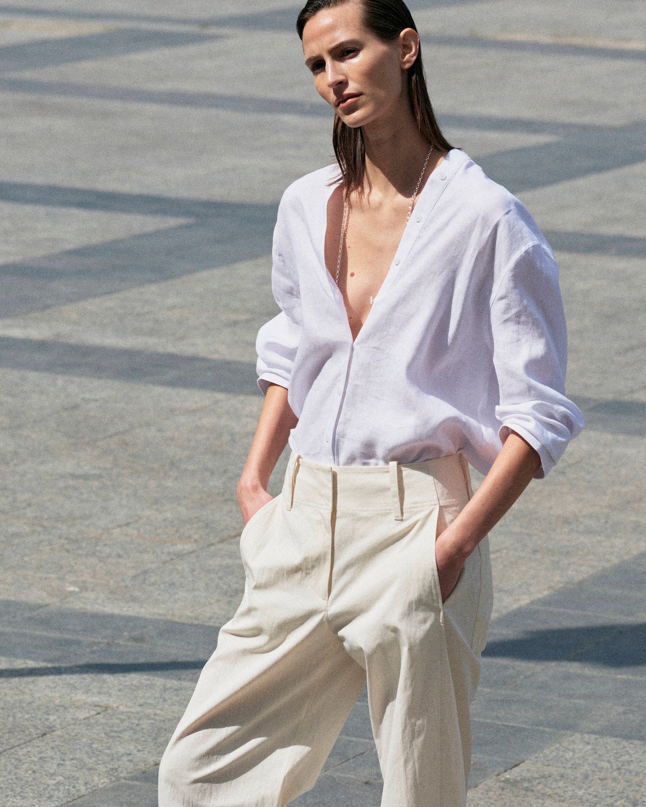 Person in a white shirt and beige pants walks outdoors on a sunny day, hands in pockets, with a minimalist stone pavement background.