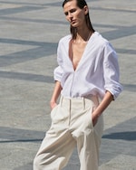 Placeholder for: Person in a white shirt and beige pants walks outdoors on a sunny day, hands in pockets, with a minimalist stone pavement background.
