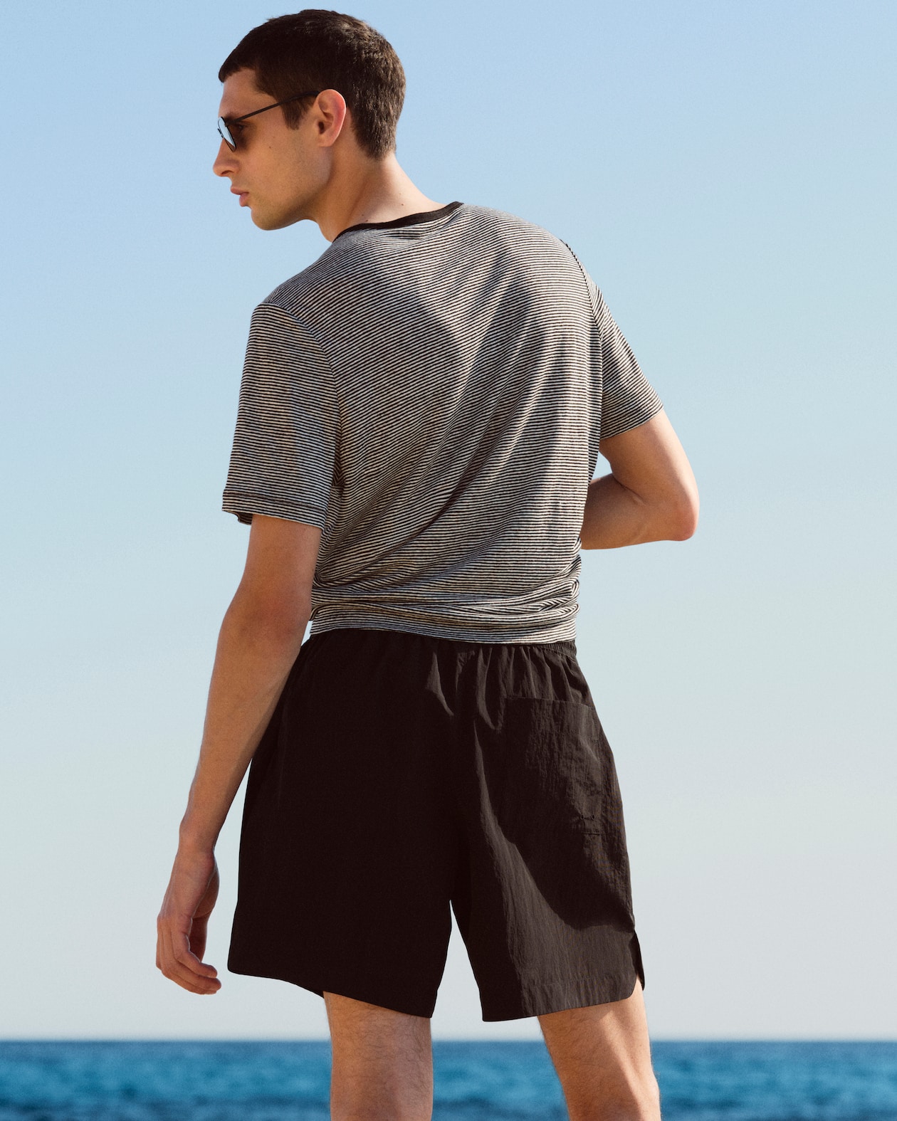 A person in sunglasses faces the sea, wearing a striped T-shirt and black shorts under a clear blue sky.