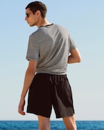 Placeholder for: A person in sunglasses faces the sea, wearing a striped T-shirt and black shorts under a clear blue sky.