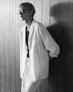 Placeholder for: Man with sunglasses in striped shirt and pants stands against wooden wall in black and white image.