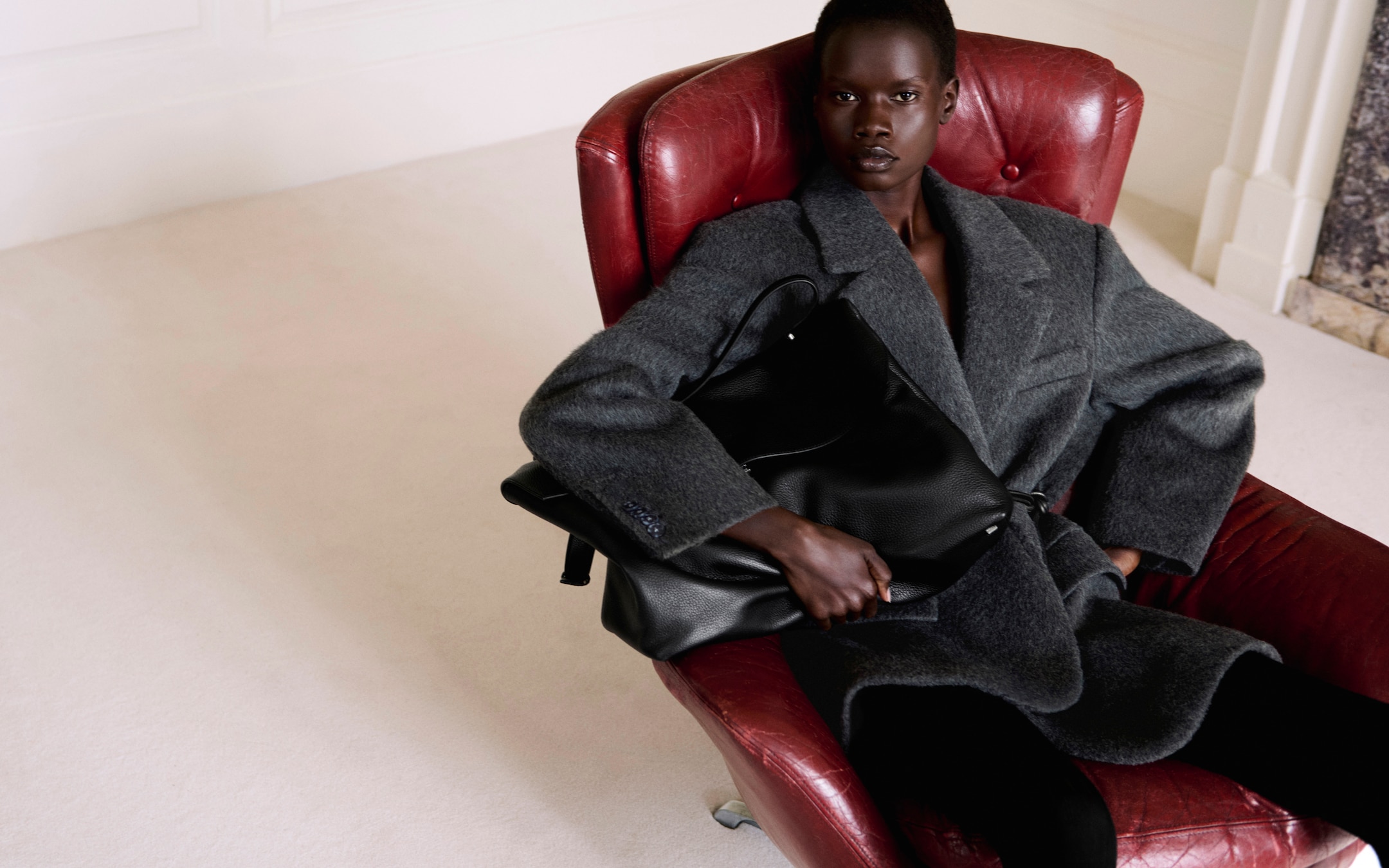 Person seated on a red leather chair wearing a gray coat, holding a large black bag, with a neutral expression and a light-colored floor background.