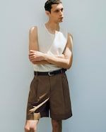 Placeholder for: A man in a white tank top and brown shorts stands with crossed arms against a light background.