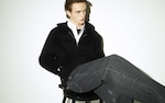 Placeholder for: Person sitting on a chair wearing a black hoodie, white shirt, and gray jeans, looking to the side against a plain background.