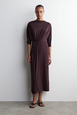 Placeholder for: Person wearing a long-sleeved, dark brown dress with gathered waist, standing against a plain white background, wearing black sandals.