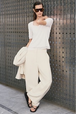 Placeholder for: Fashionable person in white outfit, sunglasses, and sandals, leaning against a metallic wall while holding a matching jacket.