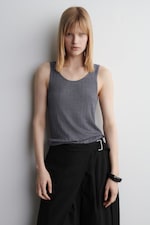 Placeholder for: Person with straight blonde hair wearing a gray sleeveless top and black pants, standing against a plain white background.