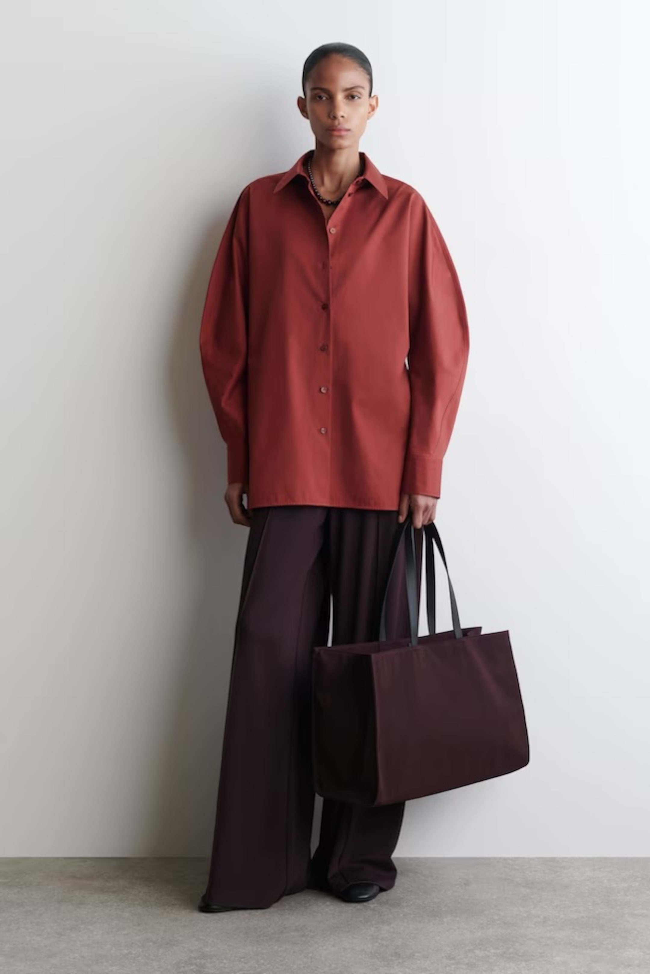 Person wearing an oversized red shirt and wide brown pants, holding a large brown tote bag against a plain white wall.