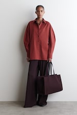 Placeholder for: Person wearing an oversized red shirt and wide brown pants, holding a large brown tote bag against a plain white wall.