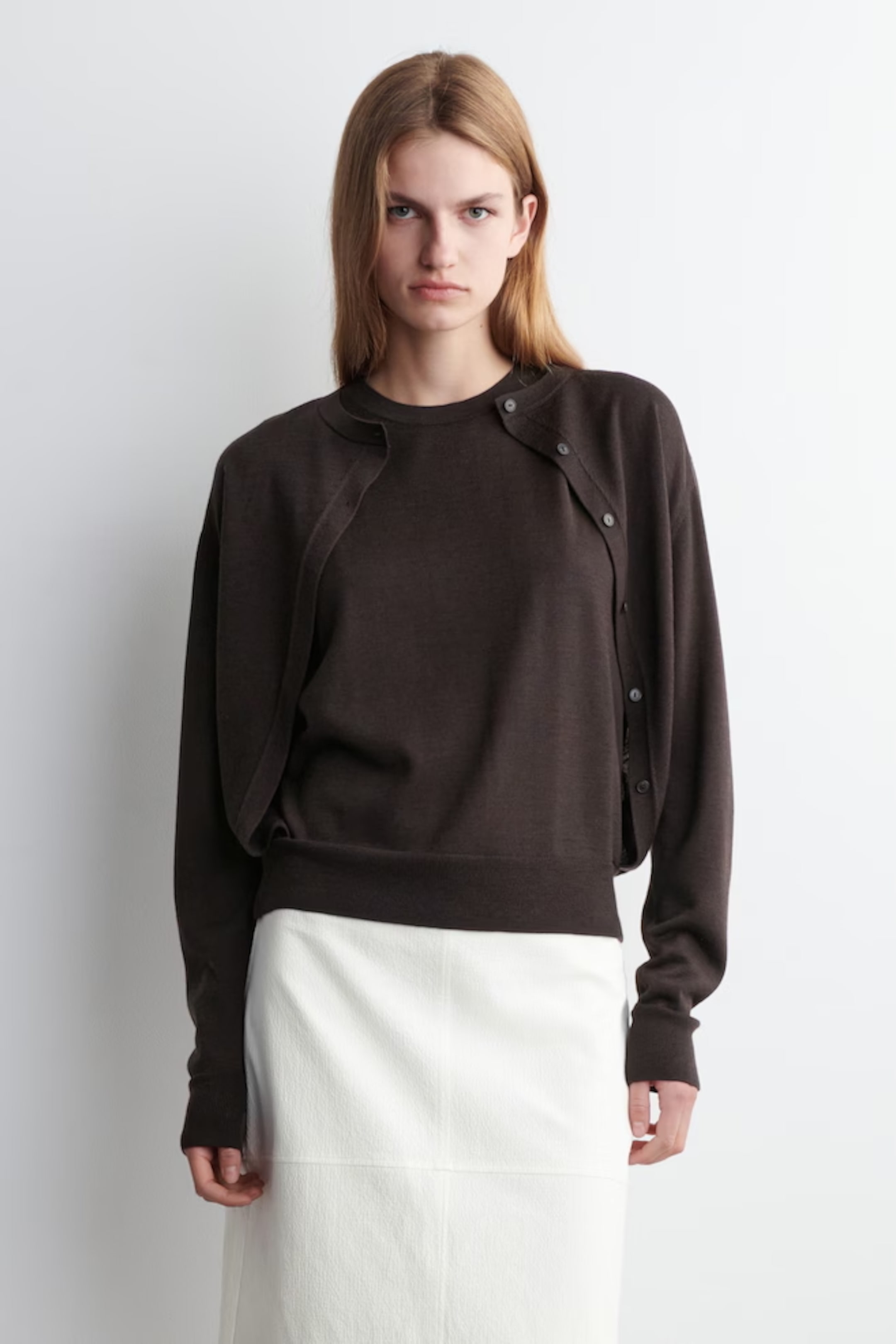 Woman in a dark sweater over a white skirt stands against a plain white background, looking directly at the camera.