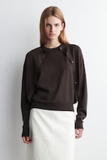 Placeholder for: Woman in a dark sweater over a white skirt stands against a plain white background, looking directly at the camera.