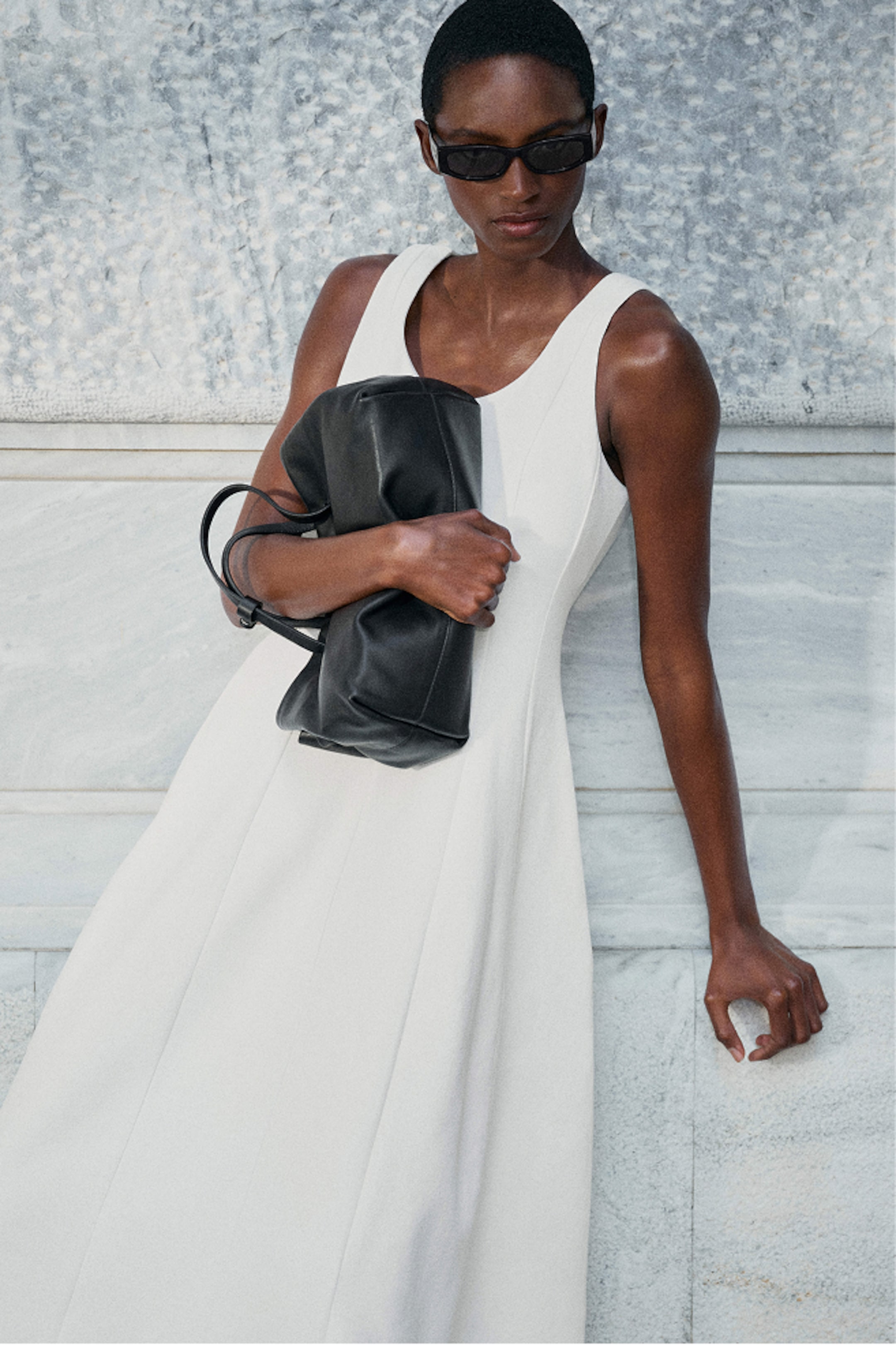 A person in a white sleeveless dress, wearing sunglasses, holds a black bag while leaning against a marble wall.