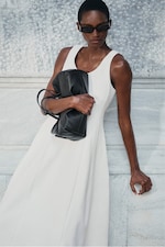 Placeholder for: A person in a white sleeveless dress, wearing sunglasses, holds a black bag while leaning against a marble wall.