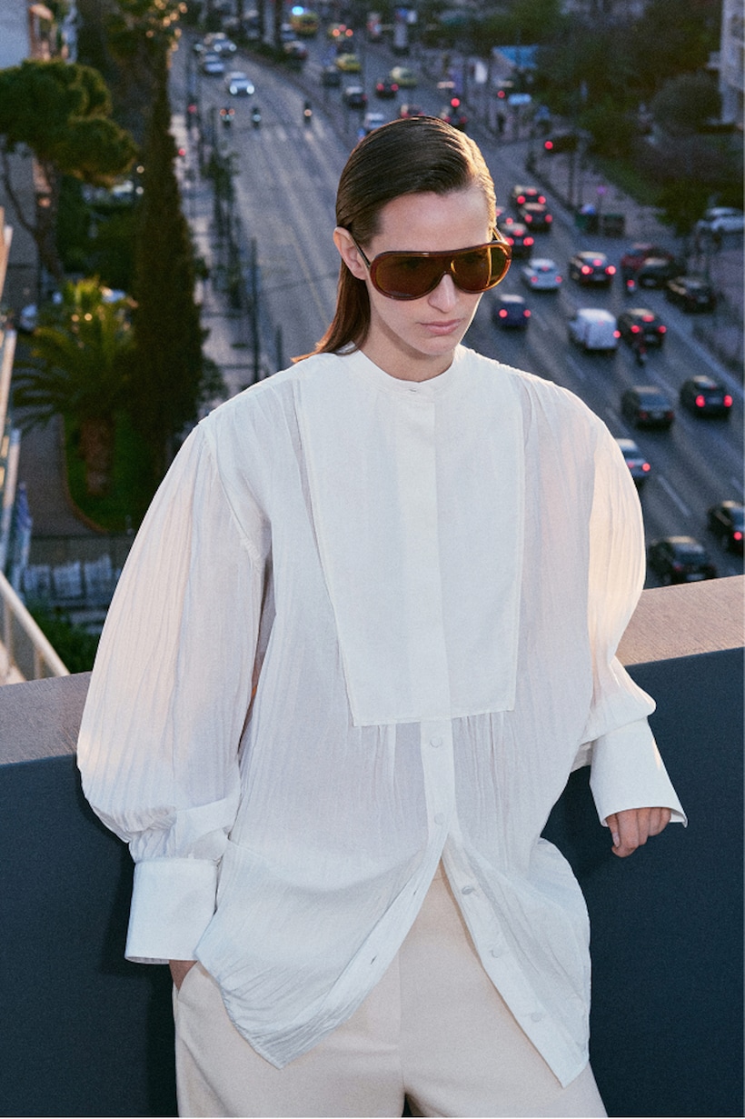 Person wearing oversized white shirt and large sunglasses, standing on a rooftop with a view of a busy city street at dusk.