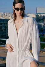 Placeholder for: Person in sunglasses wearing a white, textured outfit stands on a rooftop with a cityscape and blue sky in the background.