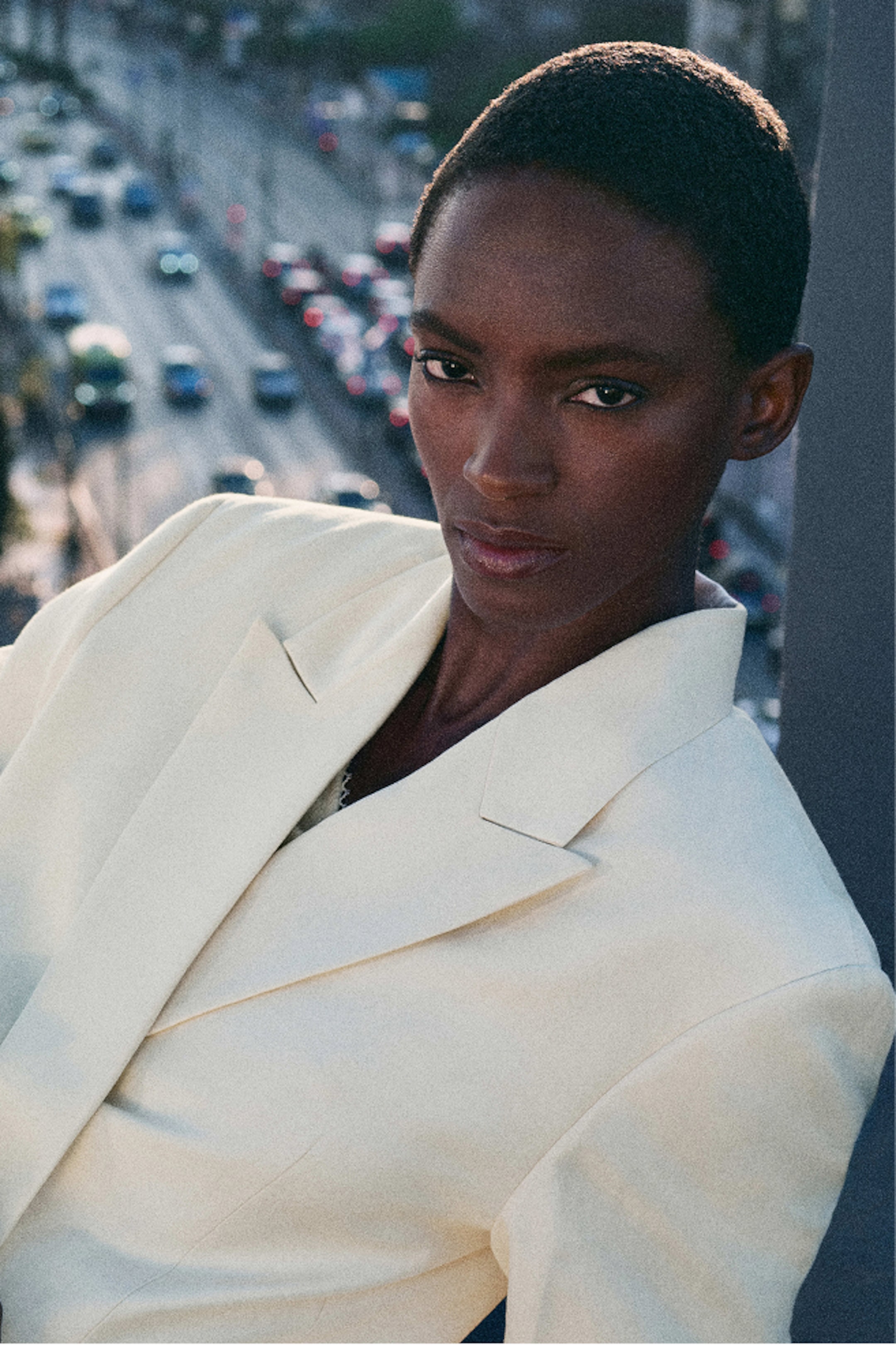 Person in a white blazer with short hair, standing outdoors with a blurred city street and traffic in the background.