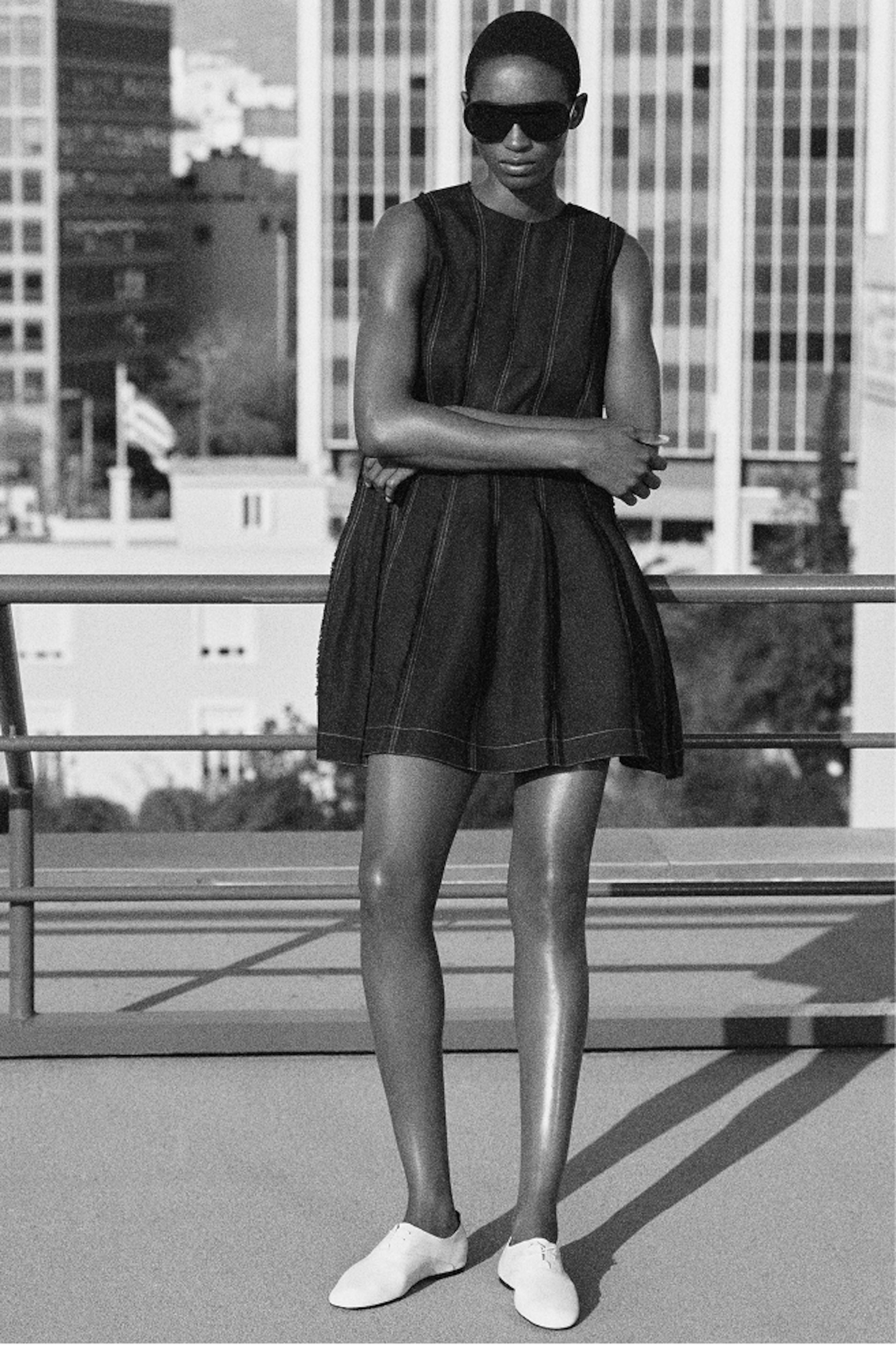 Person in a sleeveless dress and sunglasses stands on a rooftop, arms crossed, with urban buildings in the background. Black and white image.