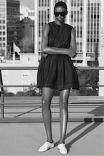 Placeholder for: Person in a sleeveless dress and sunglasses stands on a rooftop, arms crossed, with urban buildings in the background. Black and white image.