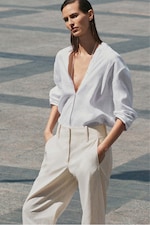 Placeholder for: Person in a relaxed pose wearing a white shirt and light cream trousers, standing outdoors on a patterned stone surface.
