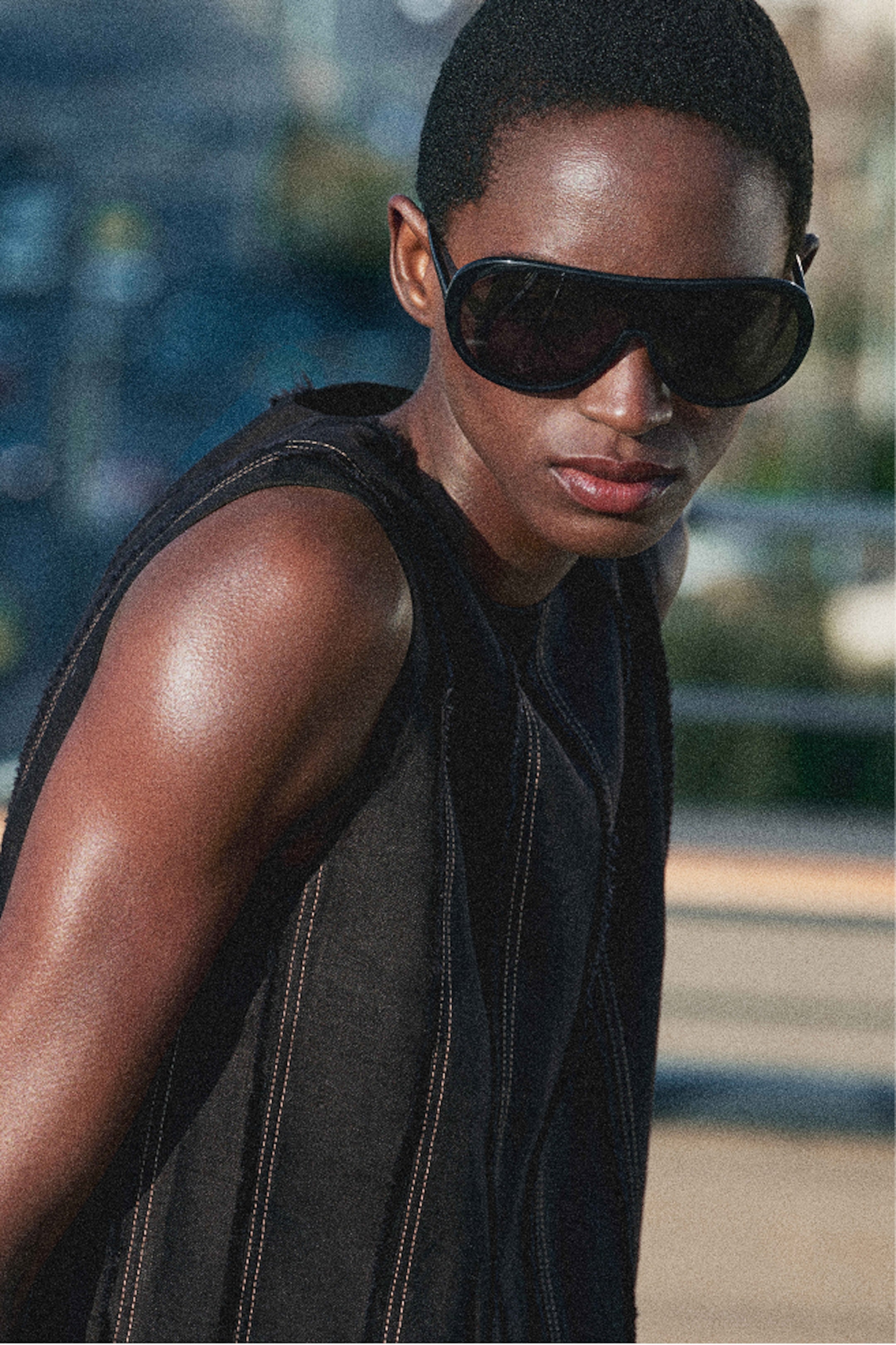 Person in dark sunglasses and sleeveless top, looking serious against a blurred outdoor background.