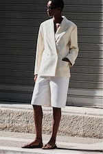 Placeholder for: Person in a light blazer and shorts stands on a street, wearing sunglasses and black sandals.