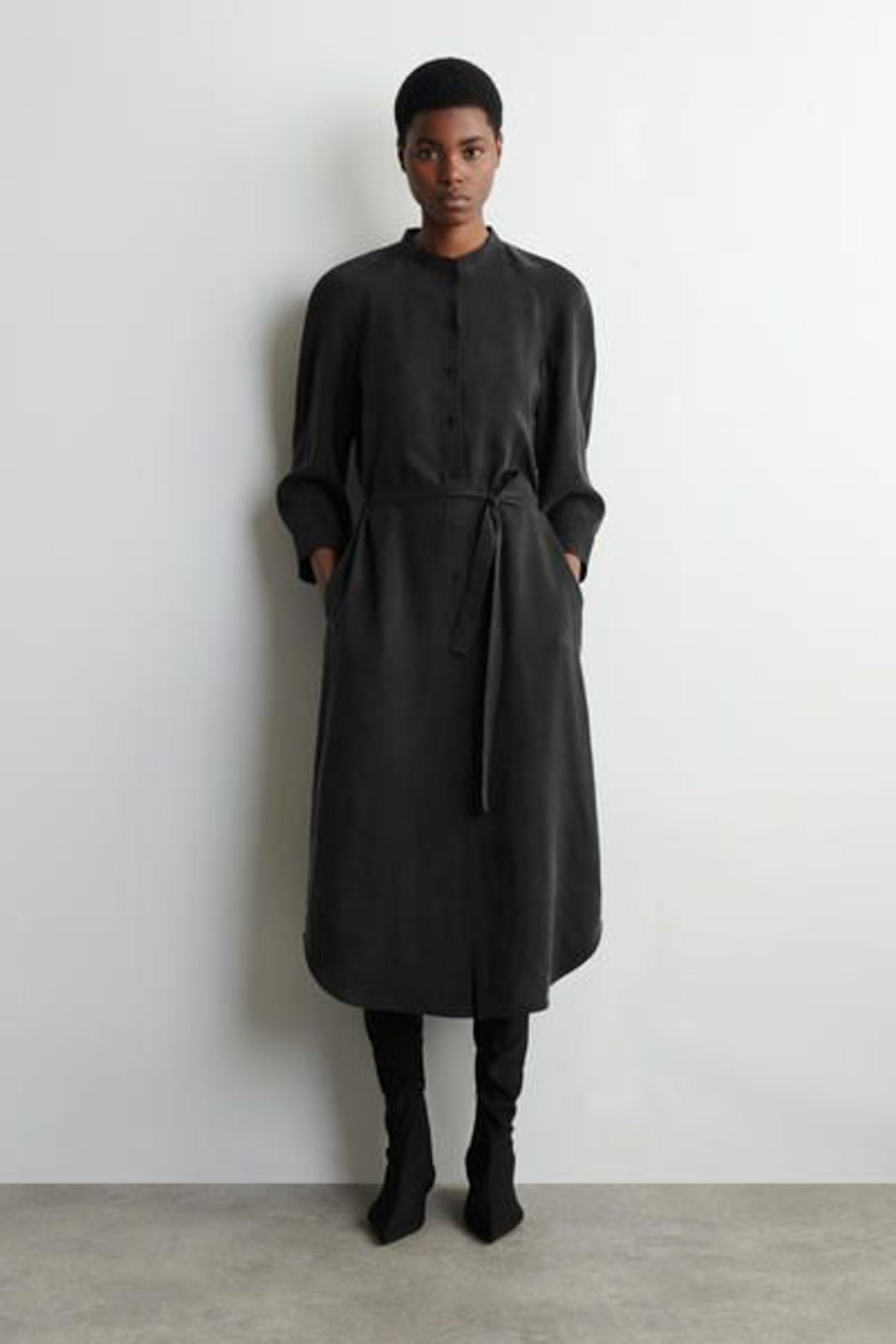 Person standing against a white wall wearing a long, black buttoned coat with pockets, a belt, and black boots.