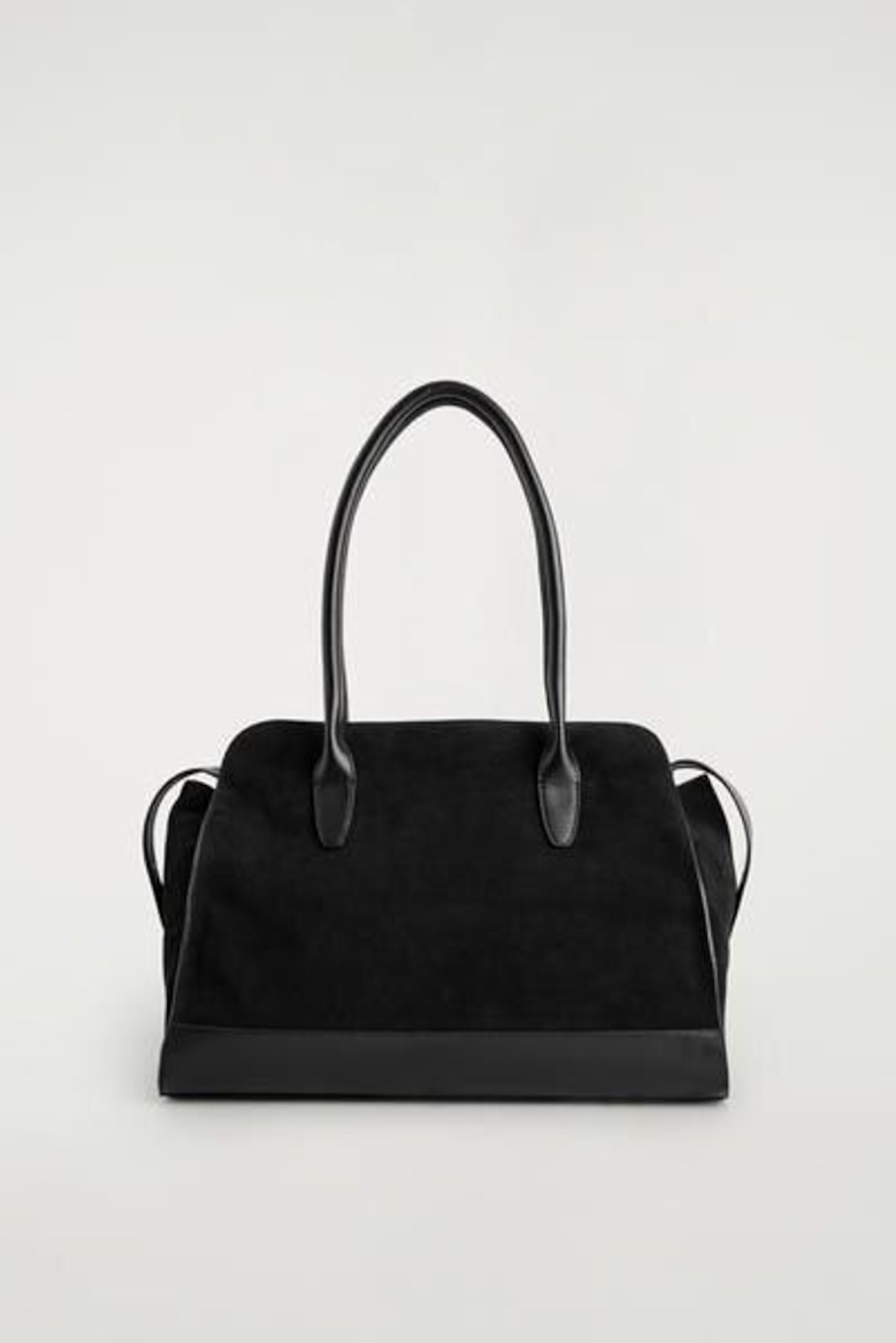 A black handbag with a structured design, suede body, and smooth leather handles, displayed against a plain gray background.