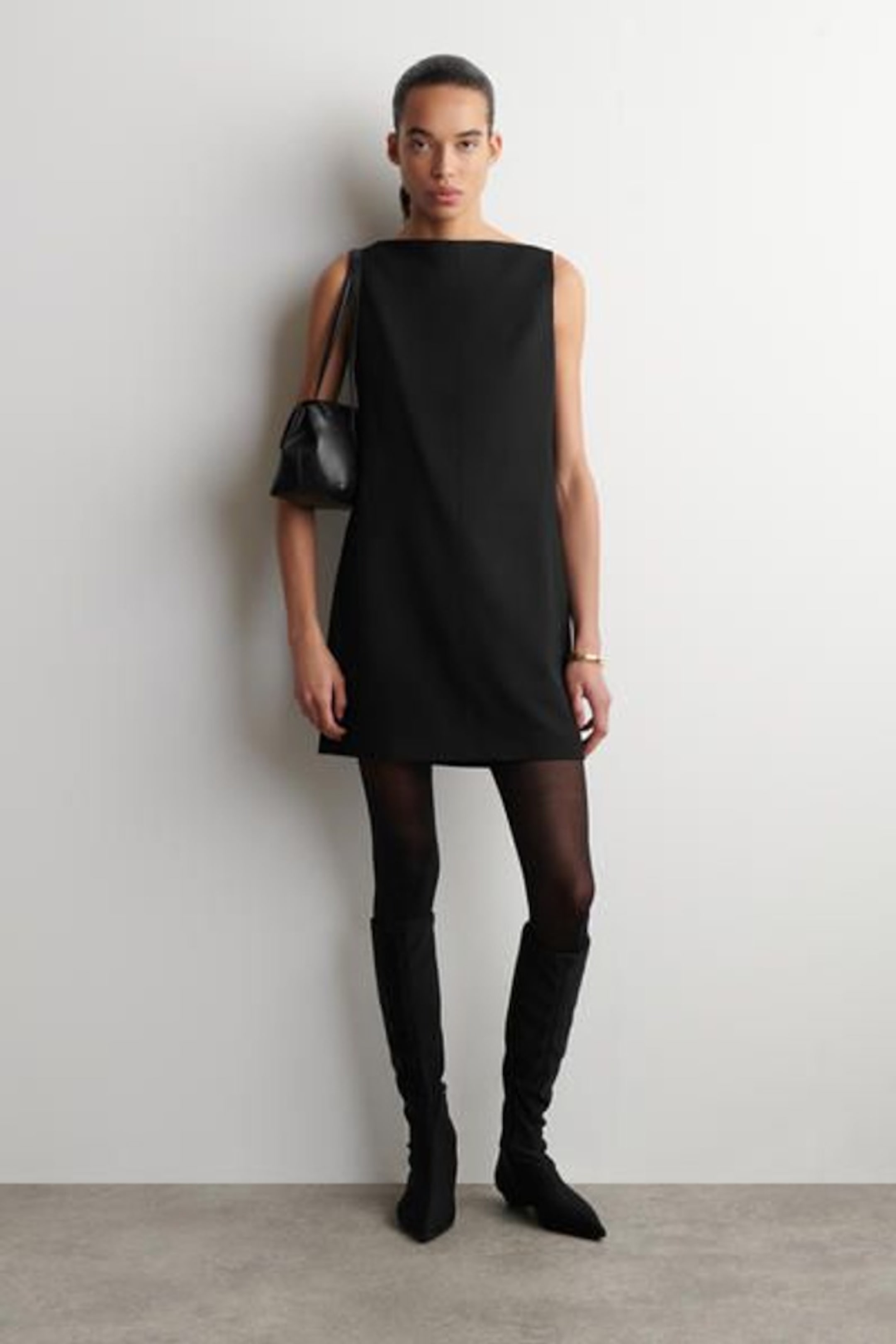 A person wearing a sleeveless black dress with knee-high black boots, holding a small black handbag, stands against a plain white background.