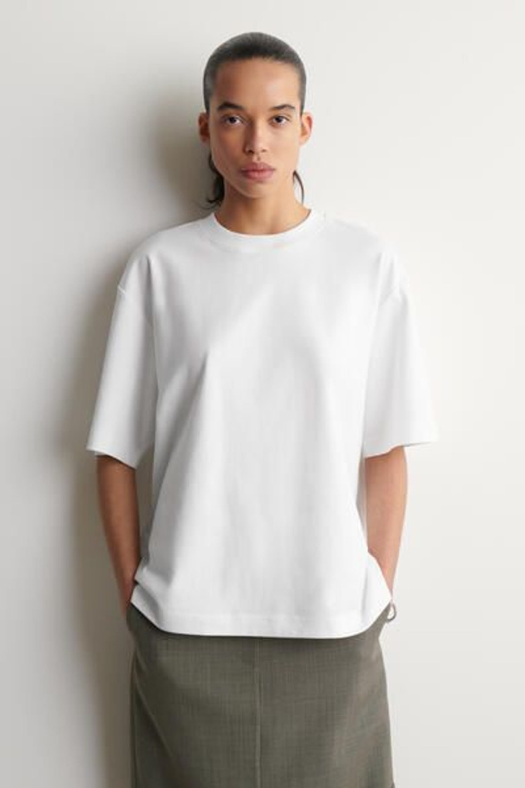 Person with hair tied back, wearing a plain white T-shirt and a gray skirt, hands in pockets, against a light-colored background.