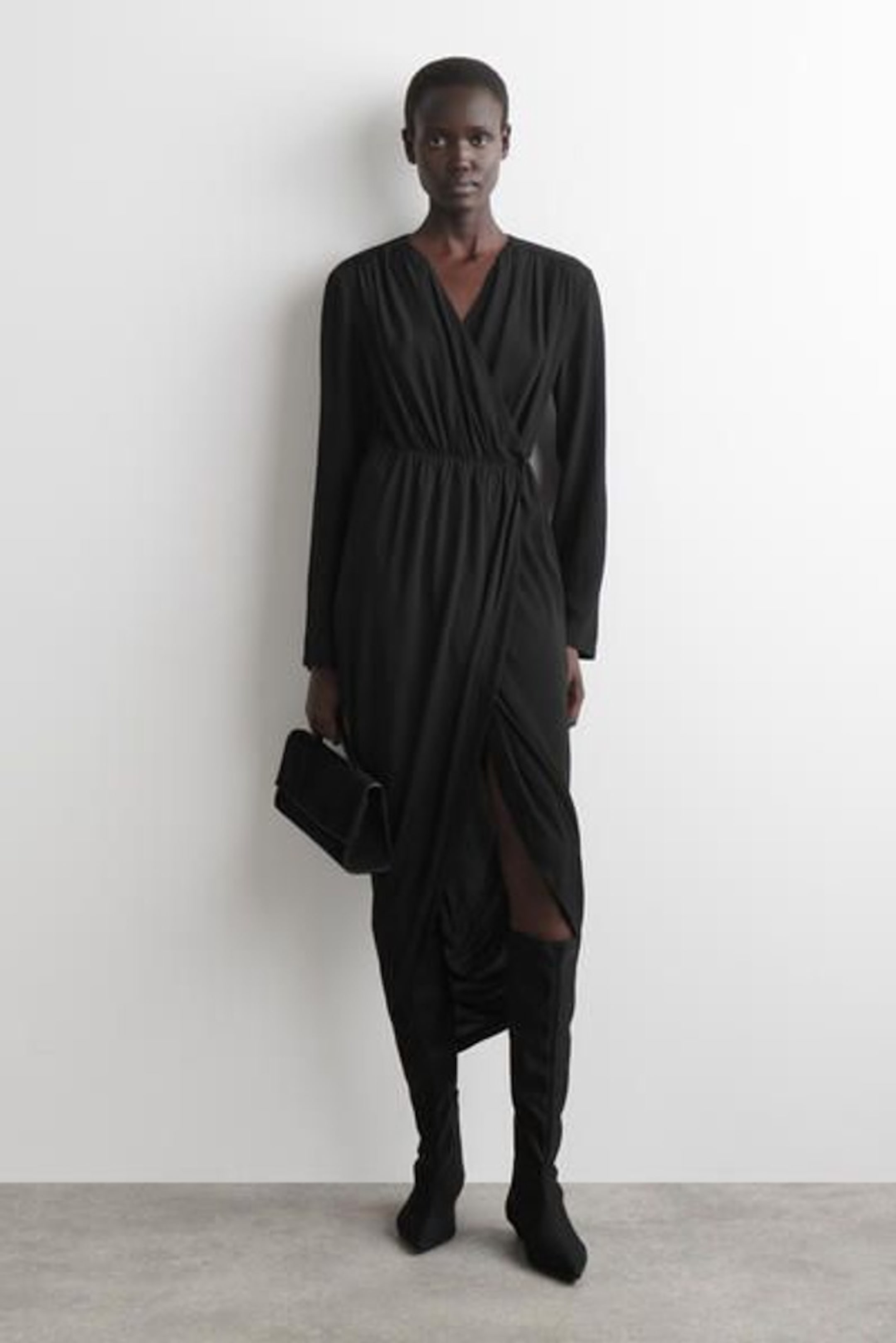 Person in a black wrap dress with long sleeves, holding a matching clutch, and wearing black knee-high boots, standing against a white background.