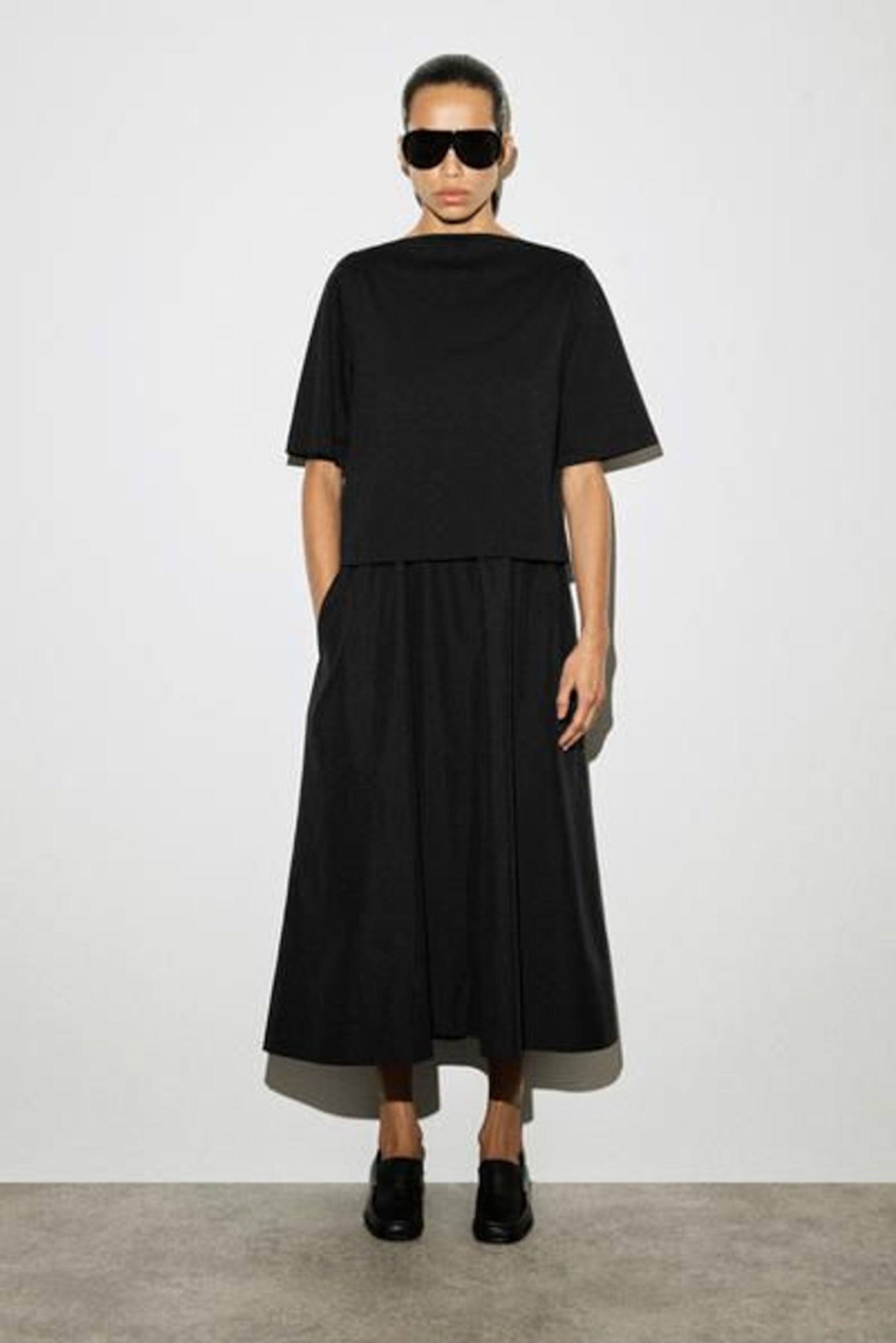 Person wearing a black outfit with wide sunglasses, standing against a plain white background.