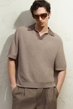 Placeholder for: Man in sunglasses wearing a beige knit polo shirt and brown pants, standing against a textured white wall with hands in pockets.