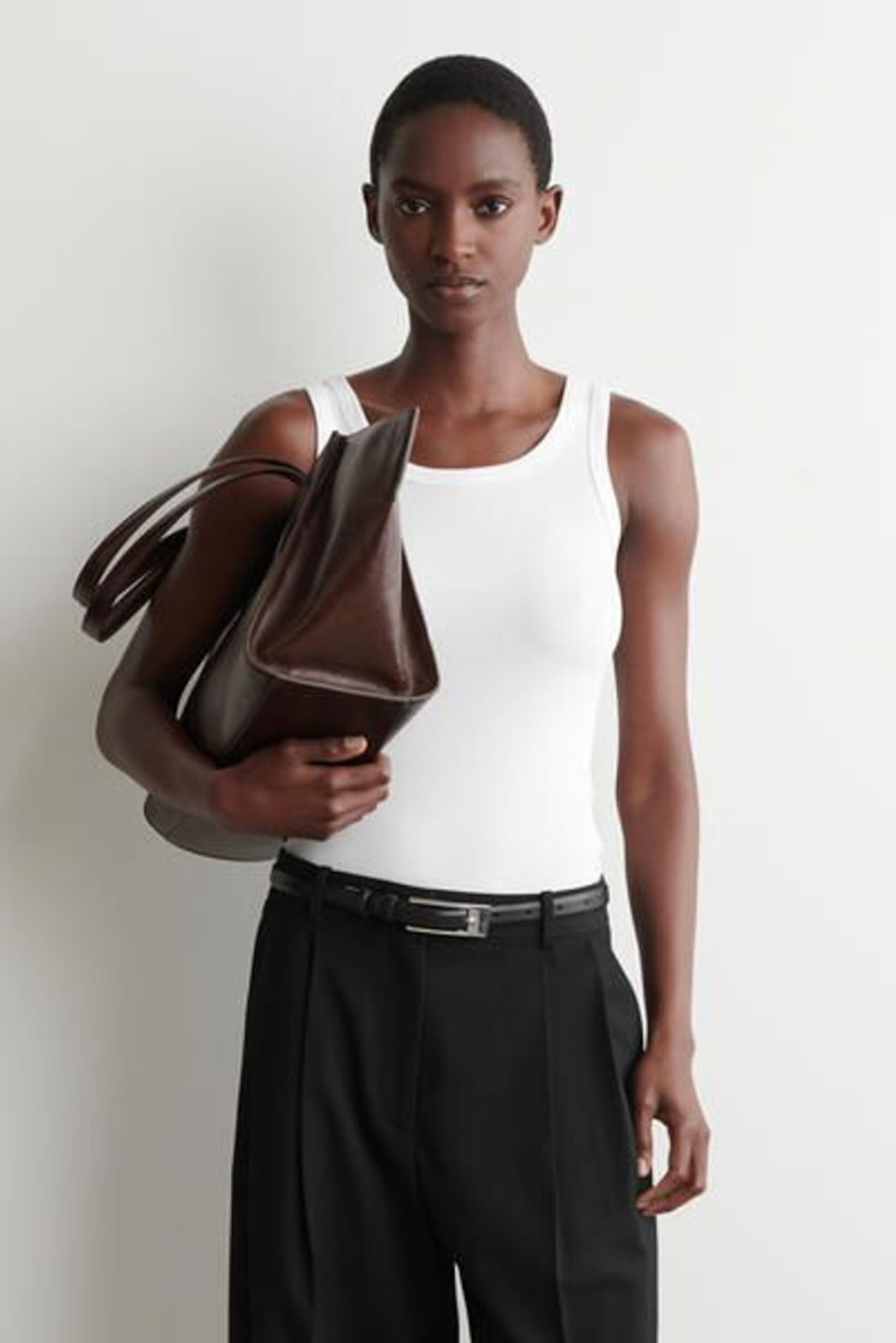 Person in a white tank top and black pants holding a large brown handbag, standing against a plain white background.