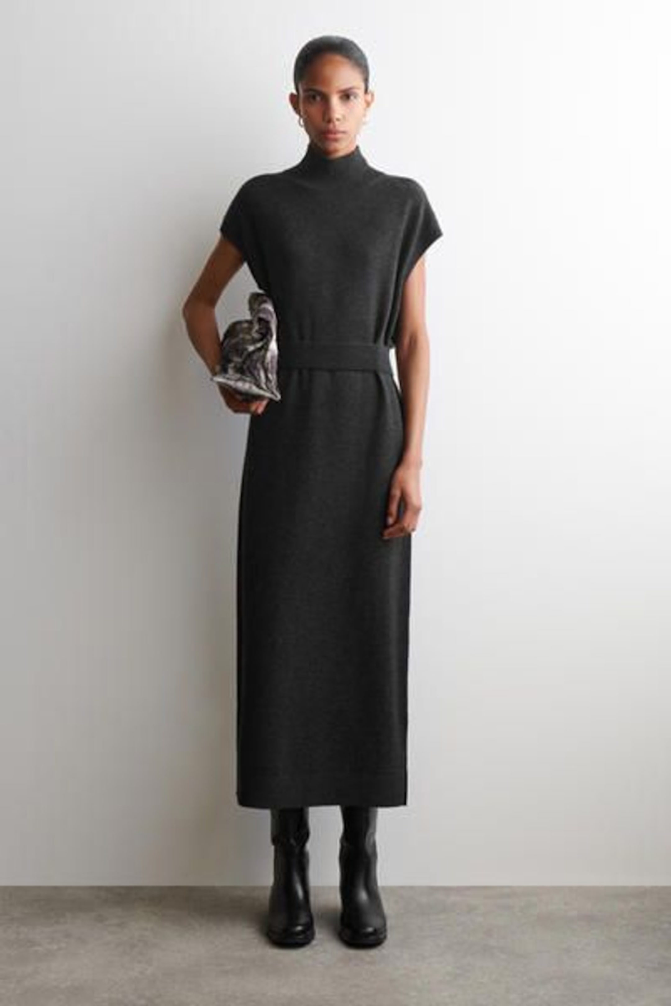 Woman in a fitted, high-neck black dress holding a patterned clutch, standing against a plain white background, wearing black boots.