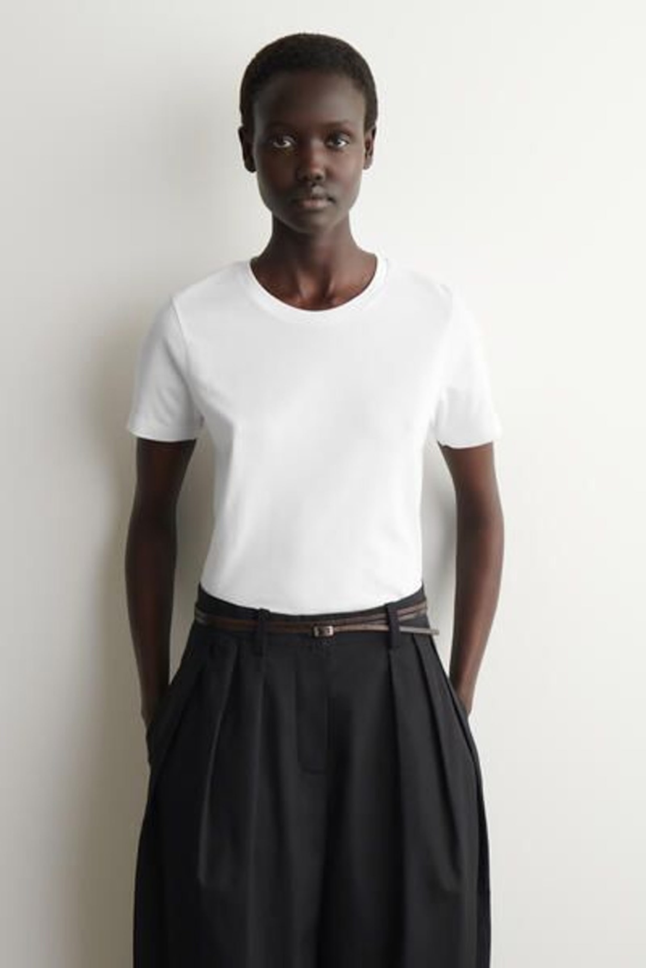 A person wearing a white T-shirt and black pleated trousers with a belt stands against a neutral wall.