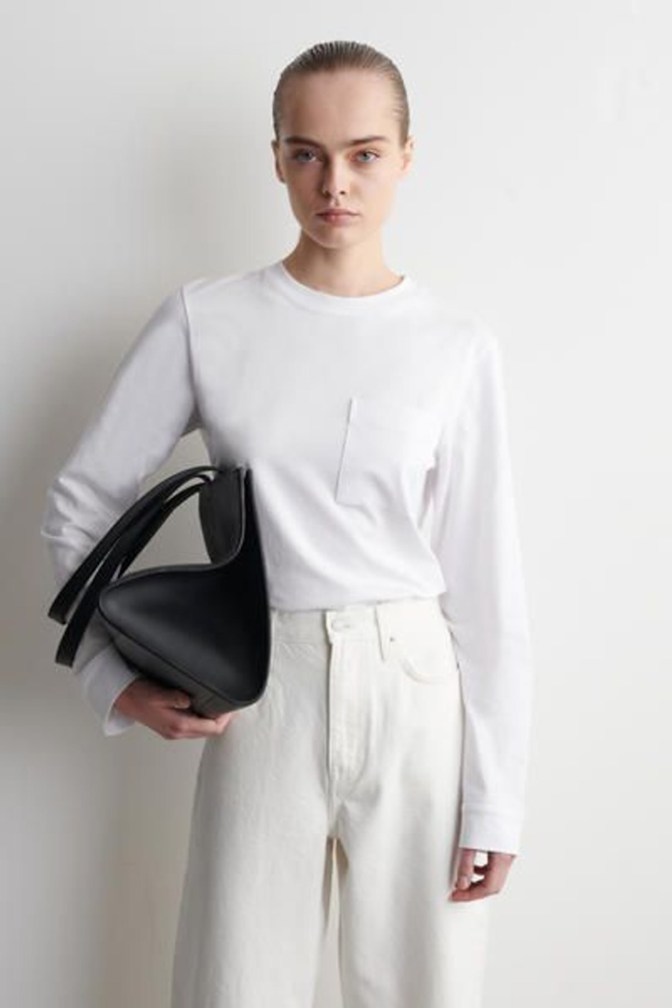 A person in a white outfit holds a large black bag against a plain background, appearing serious and stylish.
