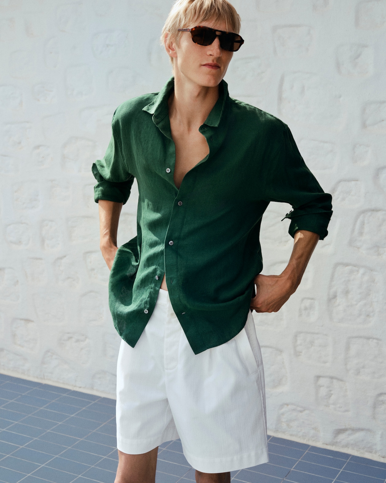Person in a green shirt and white shorts, wearing sunglasses, standing against a textured white wall and blue tiled floor.