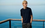 Placeholder for: A person in a navy shirt and glasses stands on a deck overlooking a calm blue sea, with hands in pockets, under a clear sky.