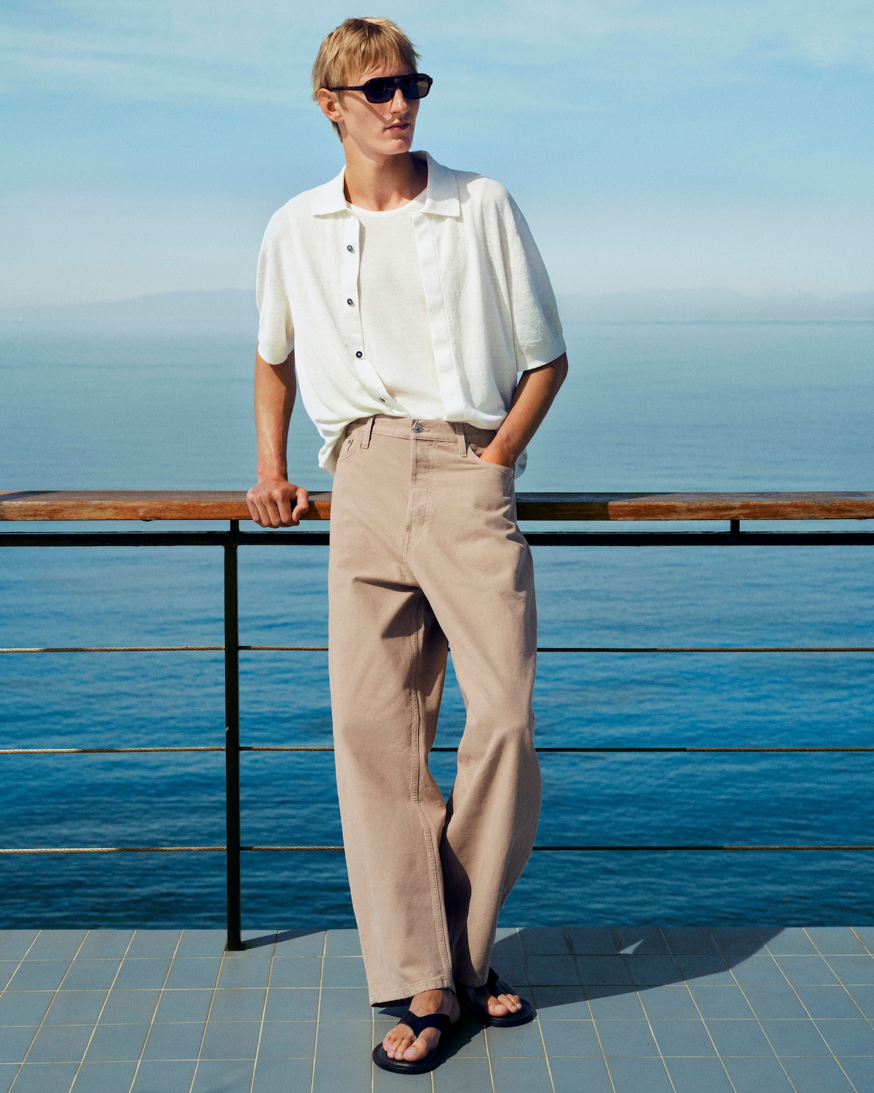 Man in sunglasses leans on a railing overlooking the sea. He's wearing a white shirt, beige pants, and sandals. Blue sky and water backdrop.