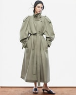 Placeholder for: A person wearing a long, belted, olive-green trench coat with high shoulders and button details, paired with black slide sandals, stands against a plain background.