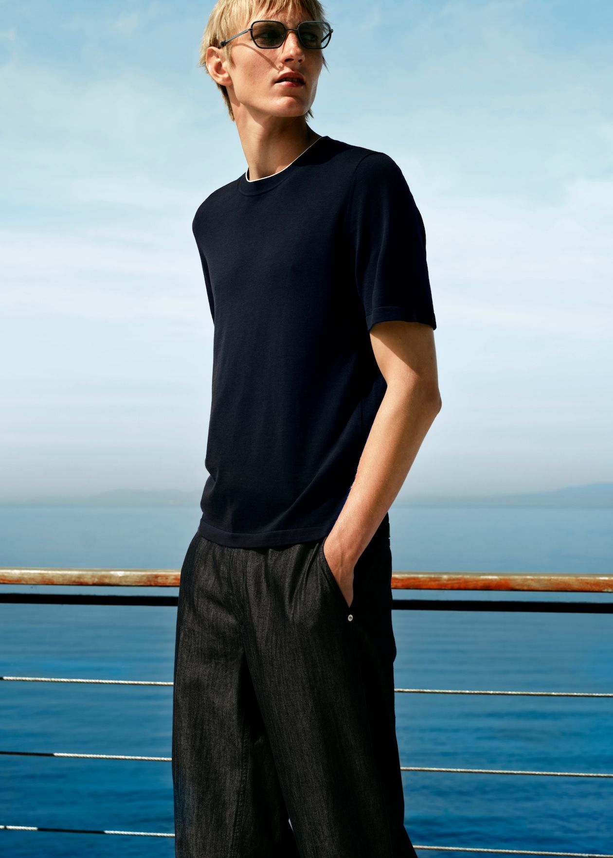 A person in sunglasses and a dark shirt stands by a railing overlooking the ocean under a clear, blue sky.