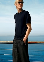 Placeholder for: A person in sunglasses and a dark shirt stands by a railing overlooking the ocean under a clear, blue sky.