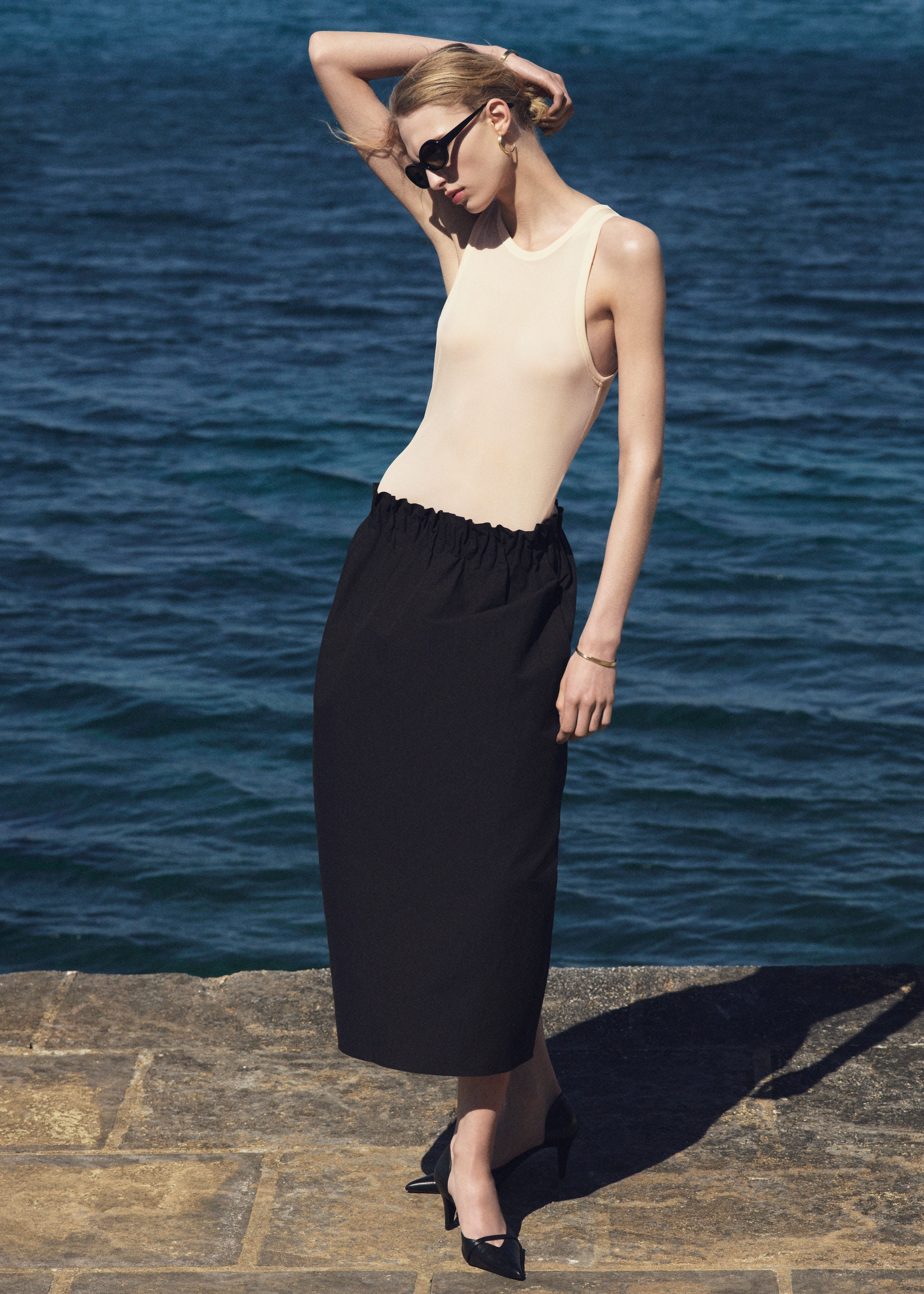 A person in sunglasses poses on a stone path by the ocean, wearing a beige sleeveless top, black skirt, and black heels, with one arm raised.