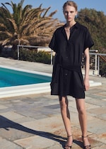Placeholder for: A person in a black dress and sandals stands by a poolside, with palm trees and blue sky in the background.
