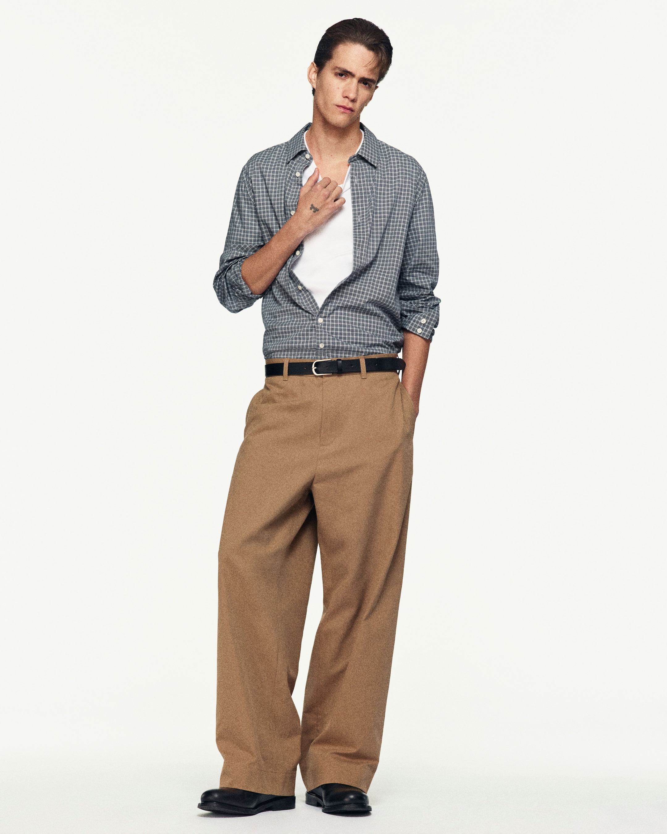 Person in a checkered shirt and brown wide-leg pants stands against a plain background, holding their collar with one hand.